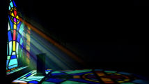 A dim old church interior lit by suns rays penetrating through a colorful stained glass window in the pattern of a crucifix reflecting colours on the floor and a speech pulpit